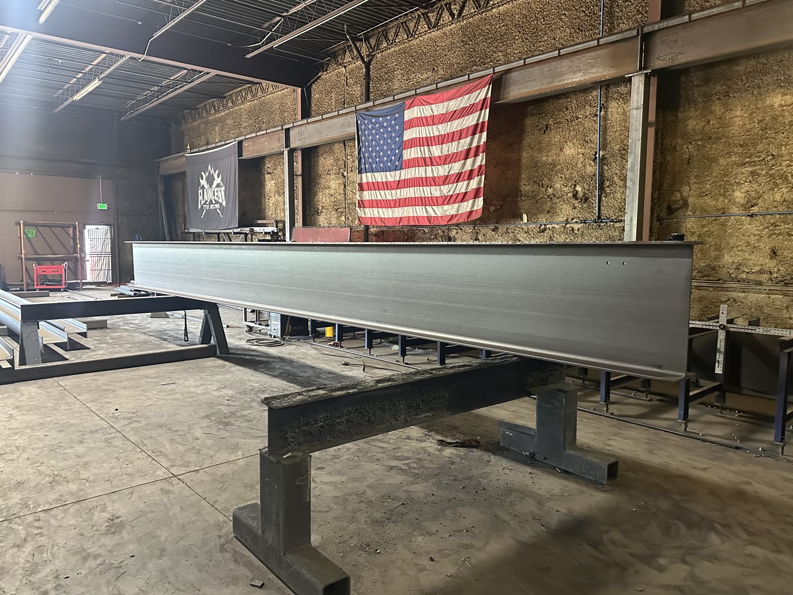 Erect-ready structural steel girder staged in the FSW shop with the American flag behind it.
