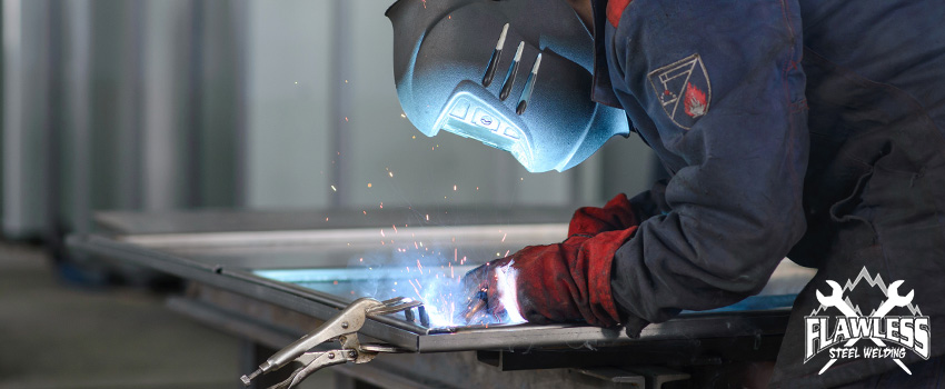 A close-up of a welder wearing a welder's hood, a blue suit, and red gloves while holding a welding torch