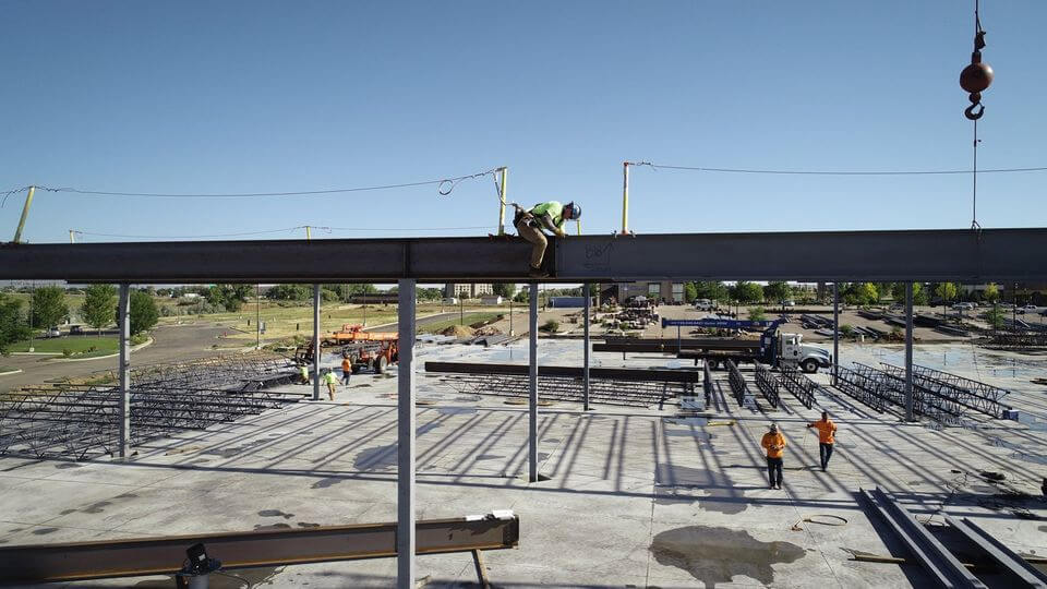 A construction worker sits on a steel beam on an ongoing construction site with tools, equipment, and co-workers in the background.